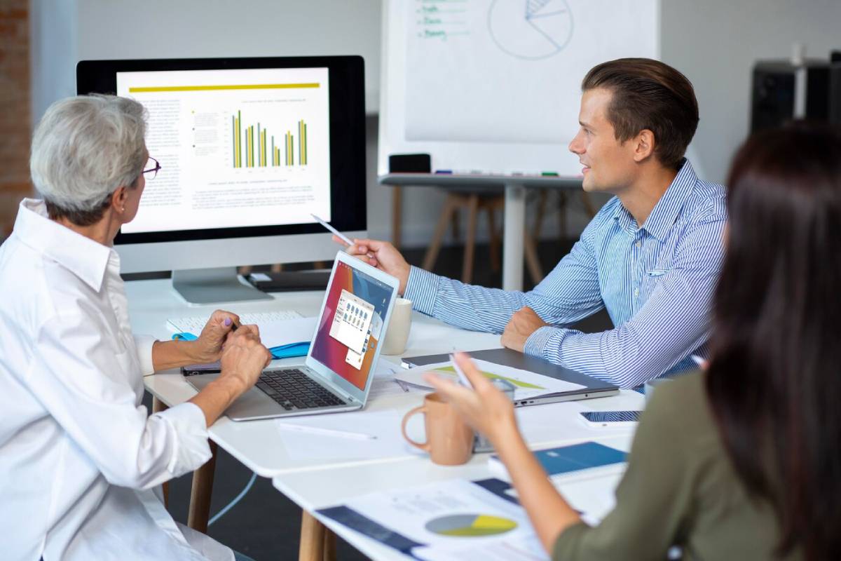 Four professionals discussing graphs and data on computers in a bright, modern office setting.