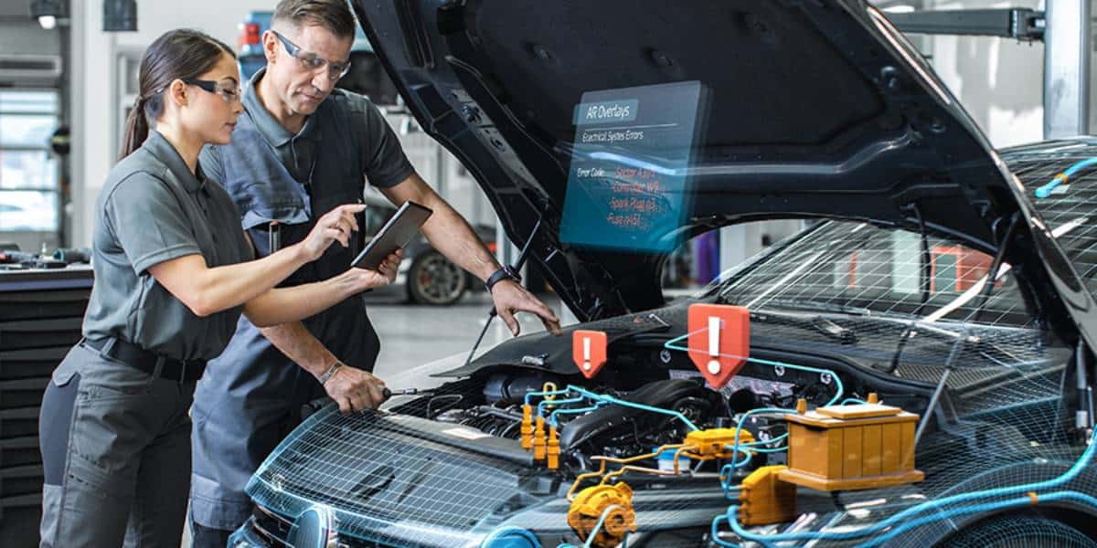 Two mechanics analyze engine diagnostics displayed on a digital screen while inspecting a car's engine compartment.