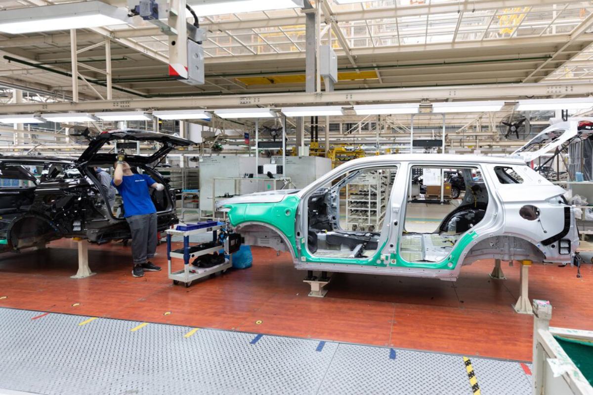 A worker assembling car frames in a factory, with partially built vehicles and equipment visible in a bright, spacious industrial environment.