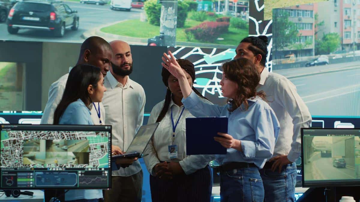 A group of professionals collaborates around computer screens displaying maps and surveillance footage in a high-tech workspace.