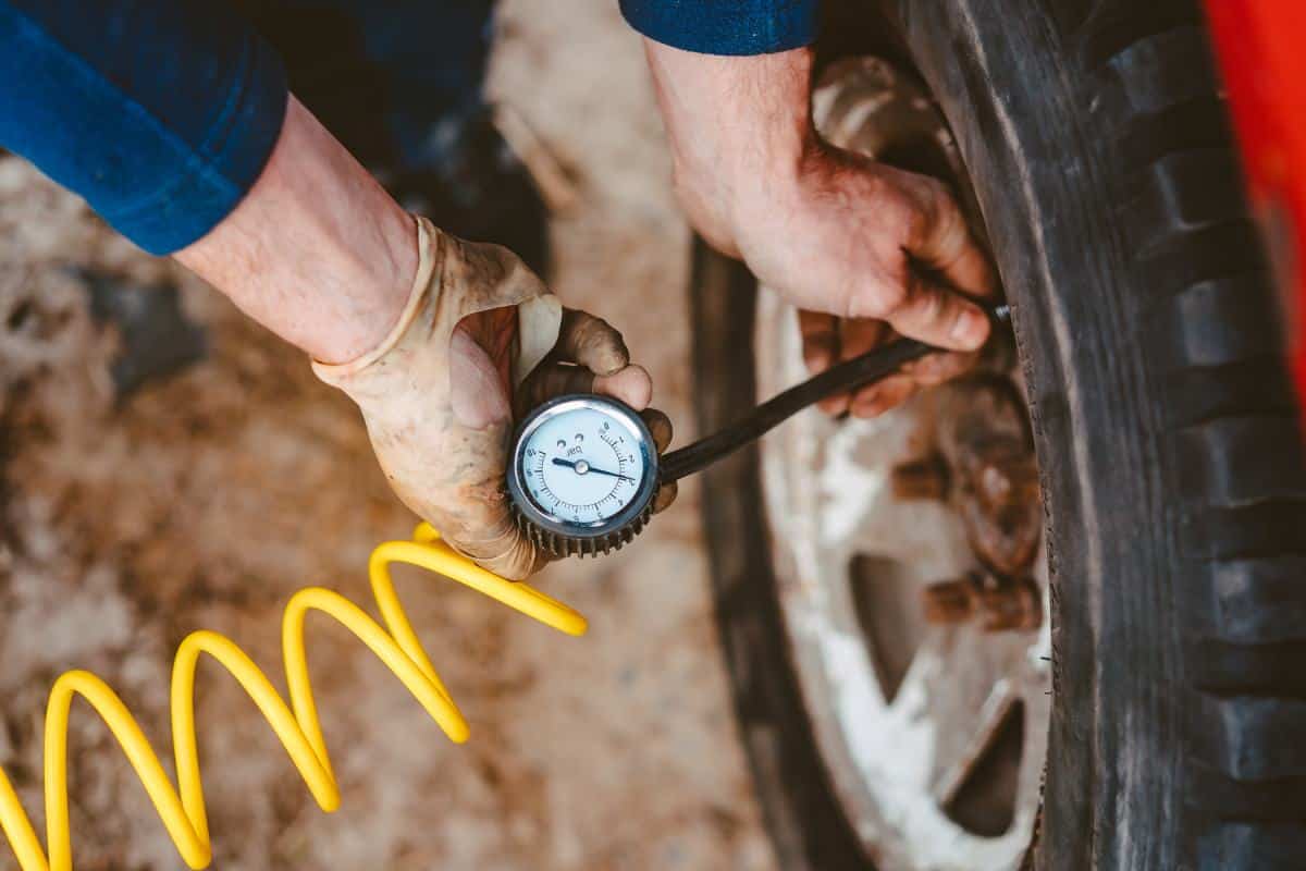 A person wearing gloves checks tire pressure with a gauge connected to an air hose, sitting beside a vehicle tire.
