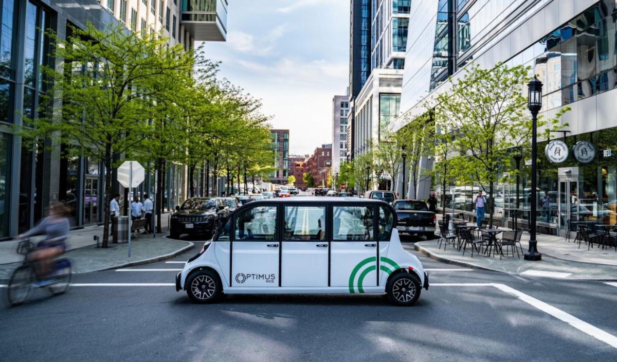 A self-driving shuttle labeled "Optimus Ride" navigates a tree-lined urban street