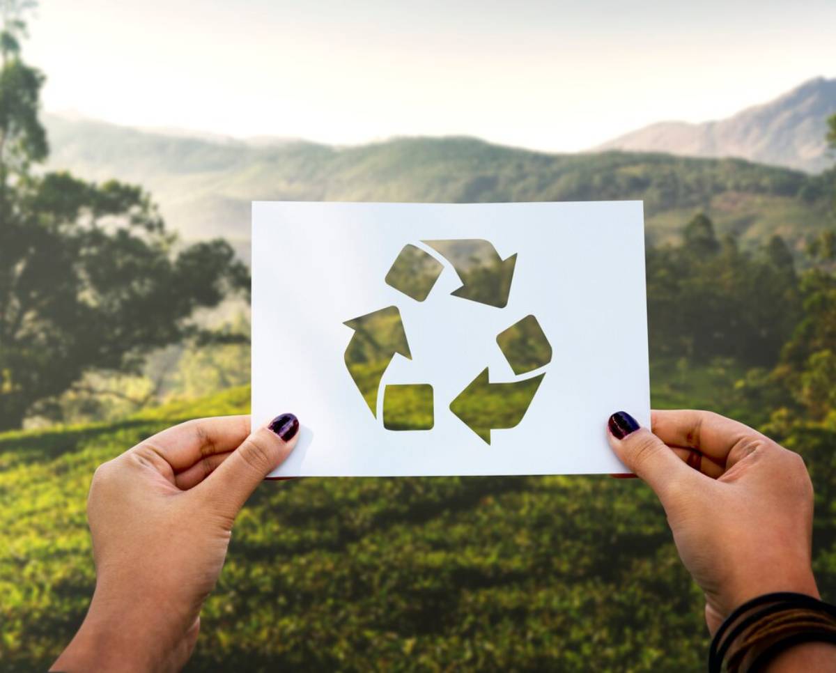 Hands holding a paper with a recycle symbol cutout, against a blurred natural landscape background.