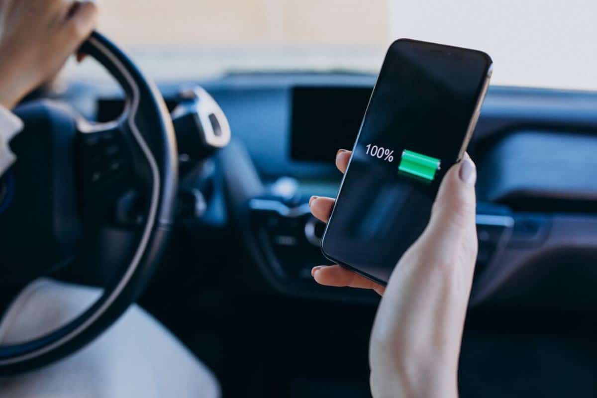 A driver's hand holds a fully charged smartphone displaying 100% battery while gripping the steering wheel of a car.