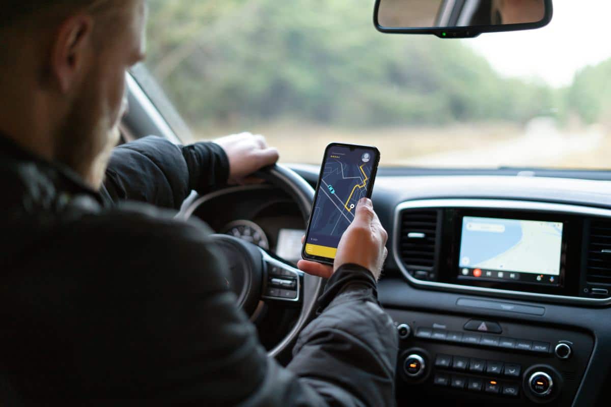 A hand with colorful rings taps a smartphone displaying a map in a car, with a steering wheel visible in the background.